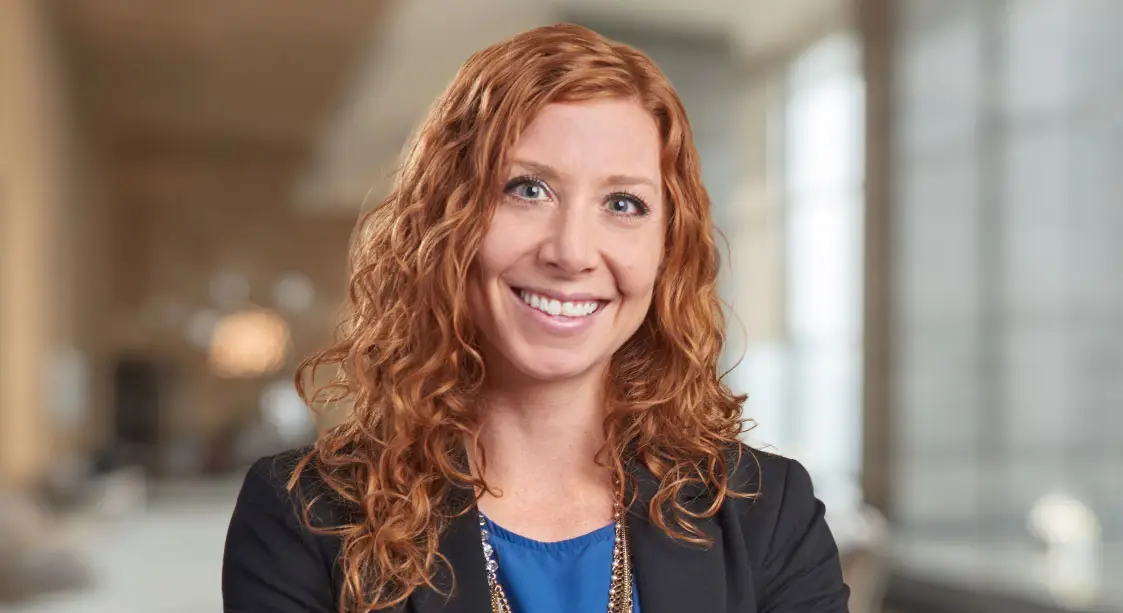A woman with long, curly red hair named Janelle Sartorio smiles She is wearing a dark blazer over a blue top and a necklace.