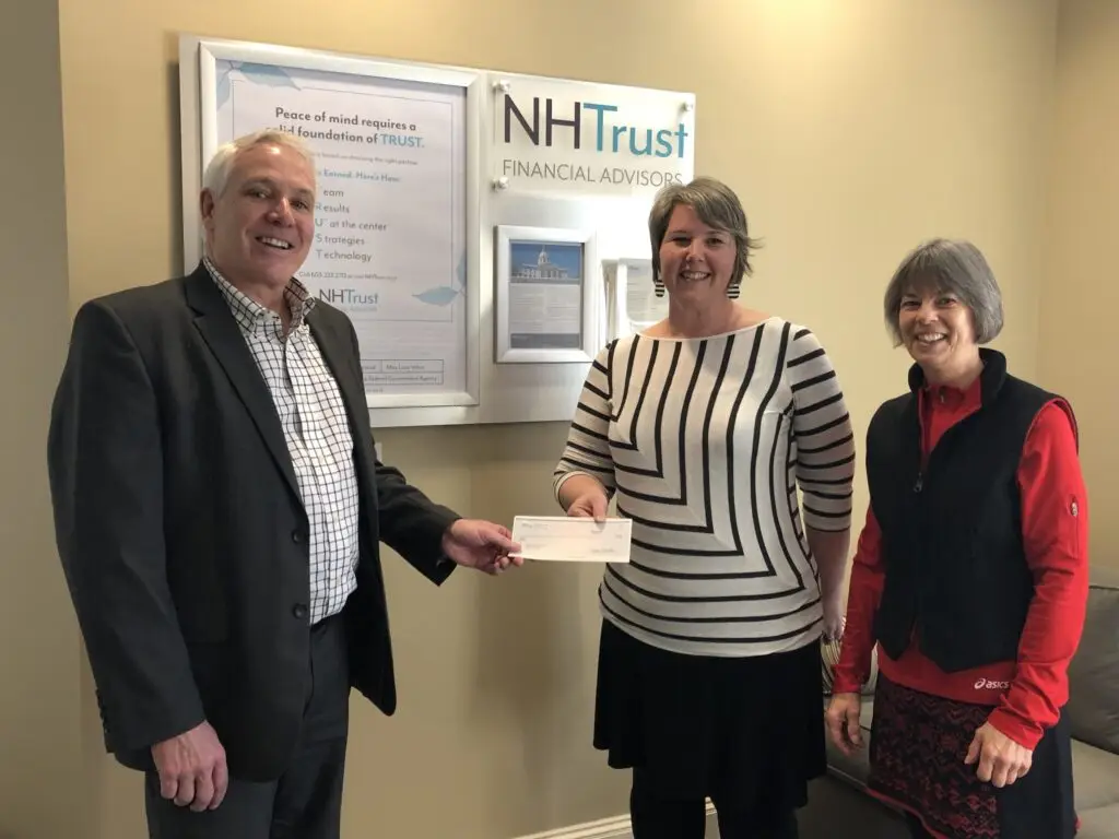 Three people smiling as one hands a check to another, standing in an office with NHTrust signage.