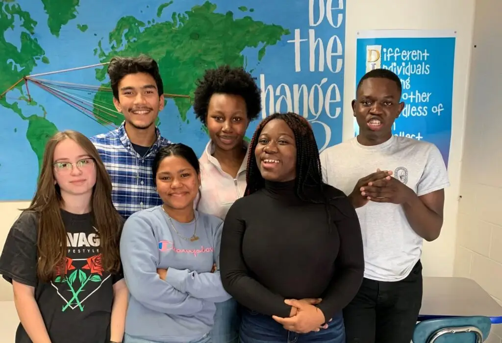 Six diverse students smiling together in a classroom, standing in front of a world map and inspirational poster.
