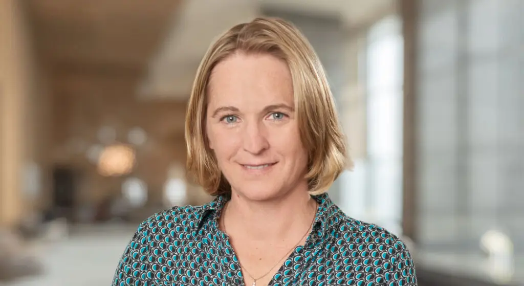 Kelly Brown, a woman with short blonde hair, wearing a teal patterned blouse, stands indoors in a softly lit room with large windows and a blurred background.