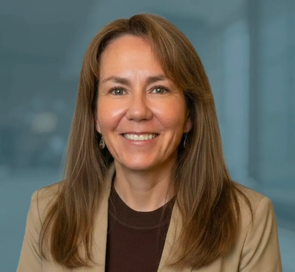 Smiling woman with long brown hair in a tan blazer, posed in front of a blurred indoor background.