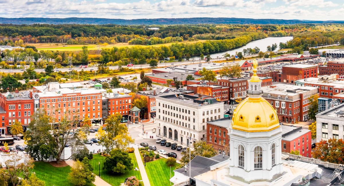 A panoramic view of Concord, New Hampshire, featuring the gold-domed New Hampshire State House, historic red-brick buildings, green trees, and a winding river in the background under a partly cloudy sky.