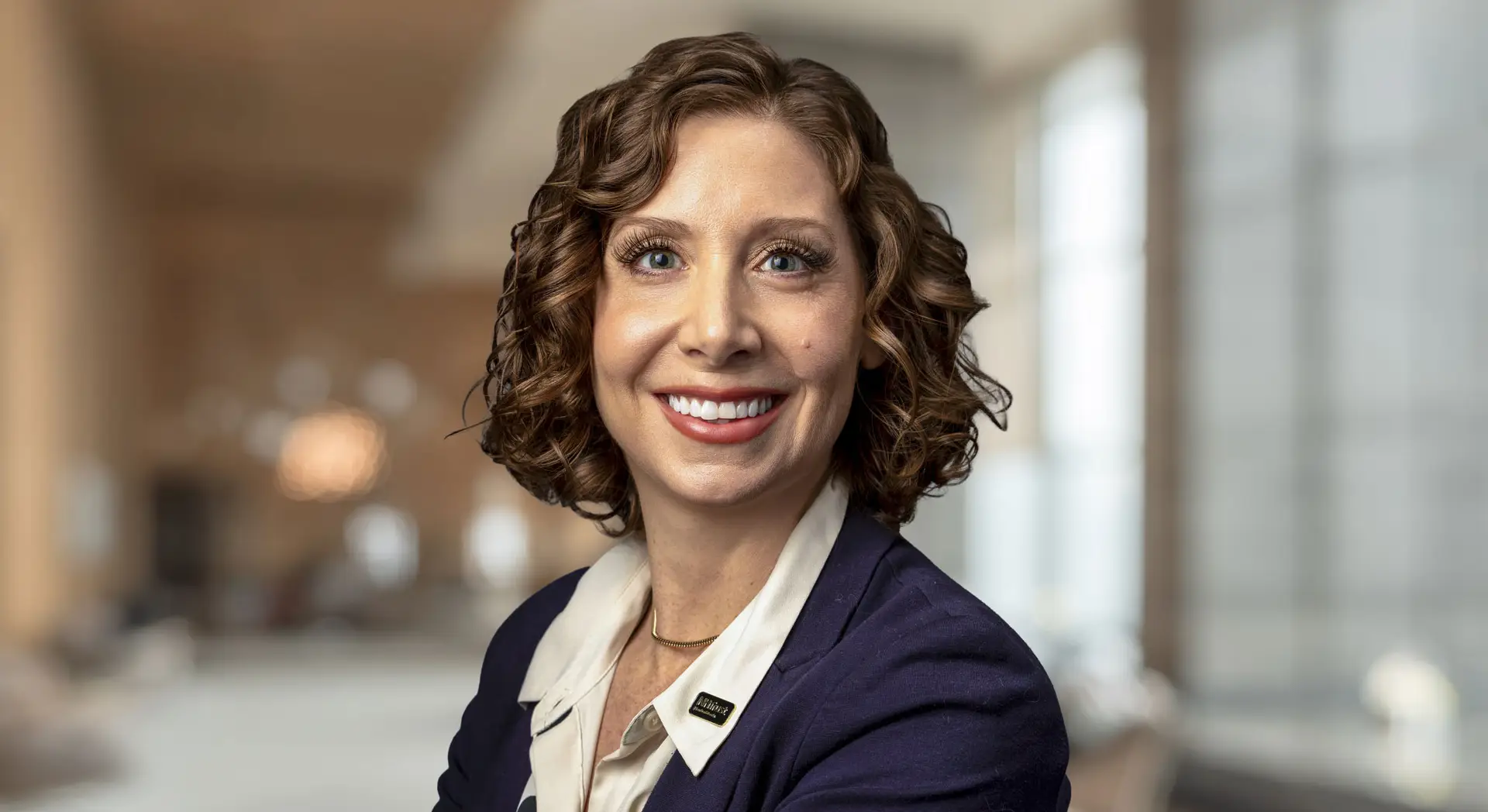 Smiling woman with curly hair in a blazer poses in a bright, blurred office setting, embodying Janelle Sartorio’s signature professional style.