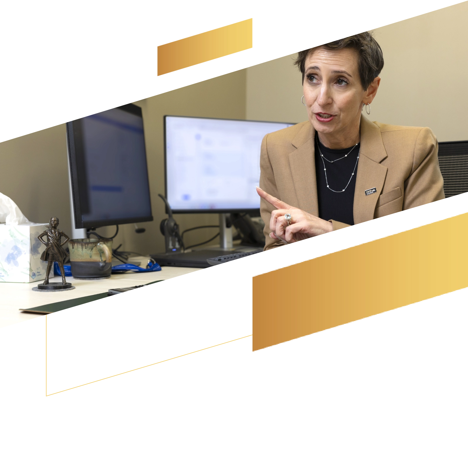 A person in a tan blazer gestures at a desk with computer monitors and a small statue.