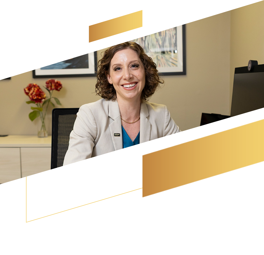 Smiling woman with curly hair in a blazer sits at a desk, with flowers and artwork in the background.