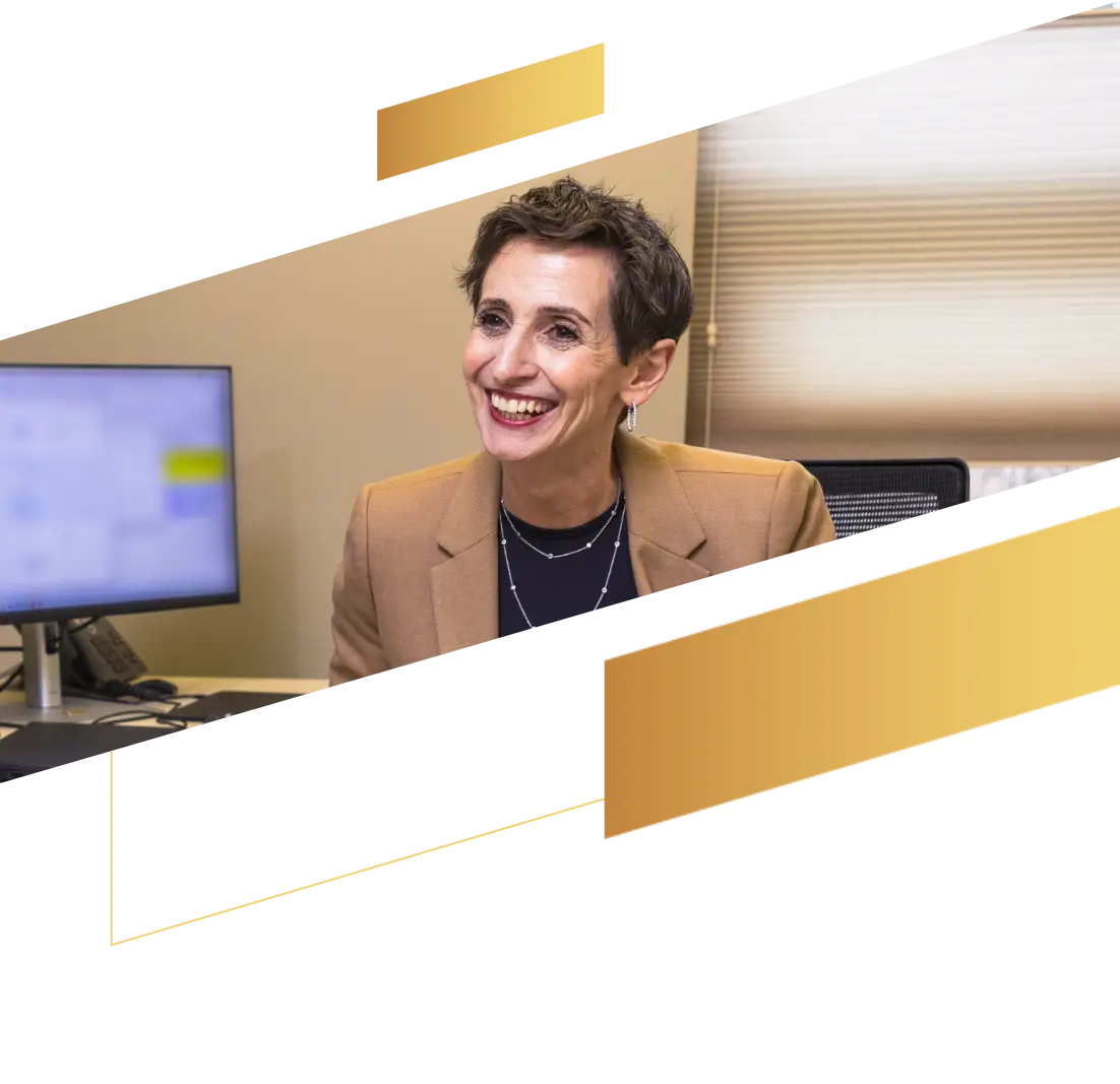 Smiling professional woman in a tan blazer sits at a desk with a computer, planning her next move with modern gold accents in the foreground.