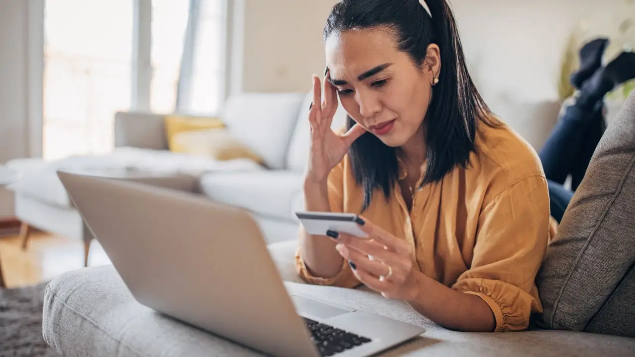 A woman sits on a couch holding a credit card in one hand and looking at a laptop screen with a concerned expression, suggesting she might be worried about an online purchase or payment.