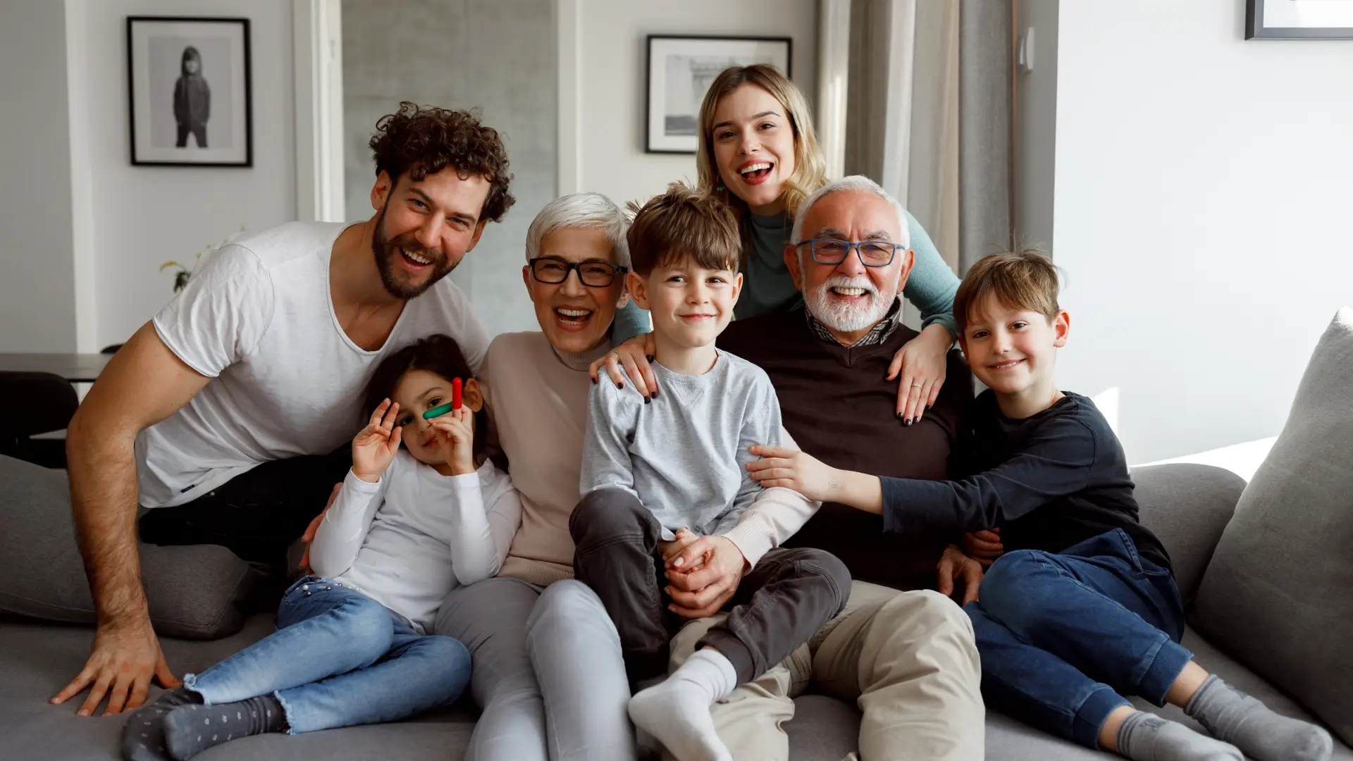 Three generations of a happy family sit close together, smiling on a couch in a bright living room.
