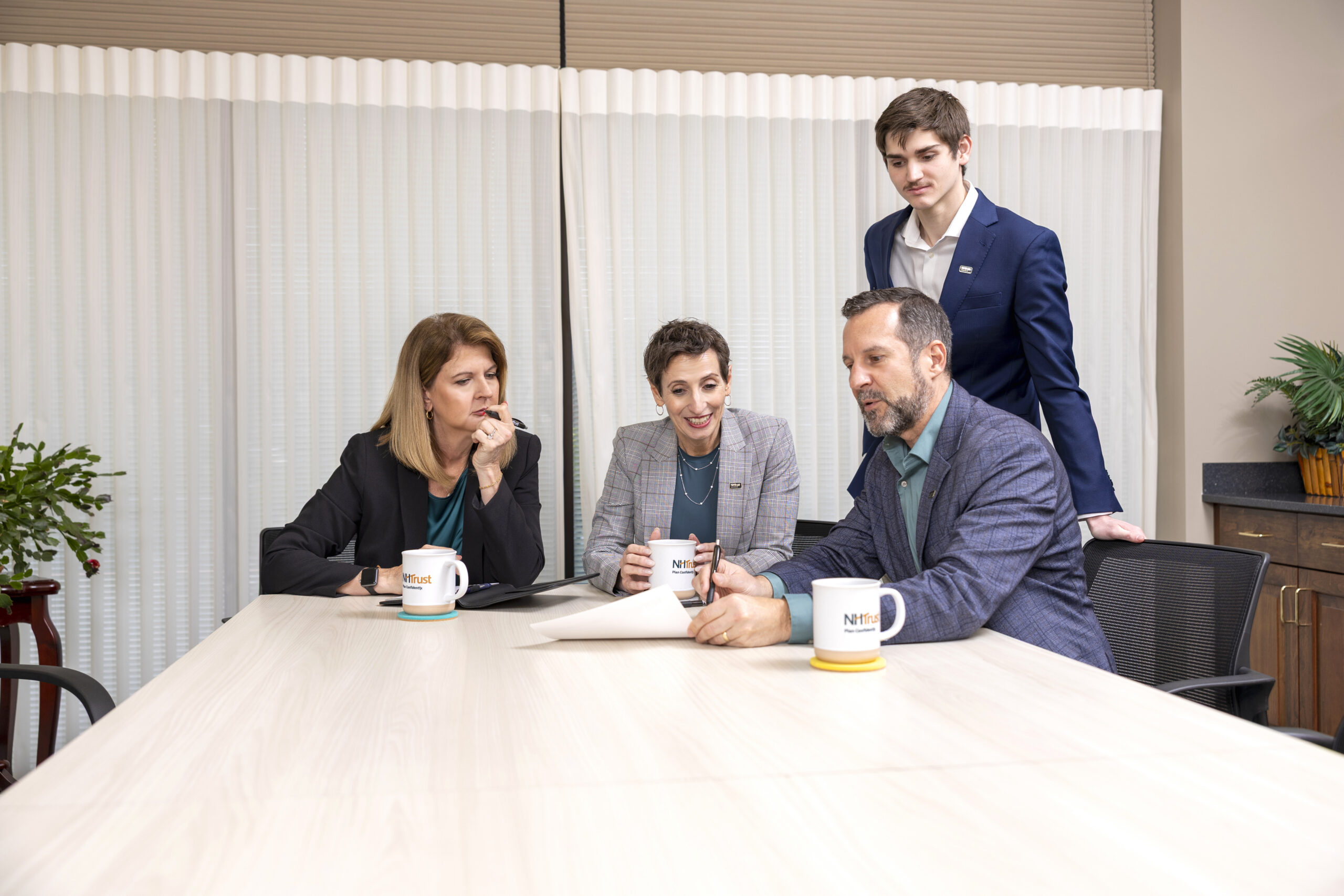 Four people in business attire sit and stand around a table, engaged in a review meeting as they discuss documents and sip coffee.