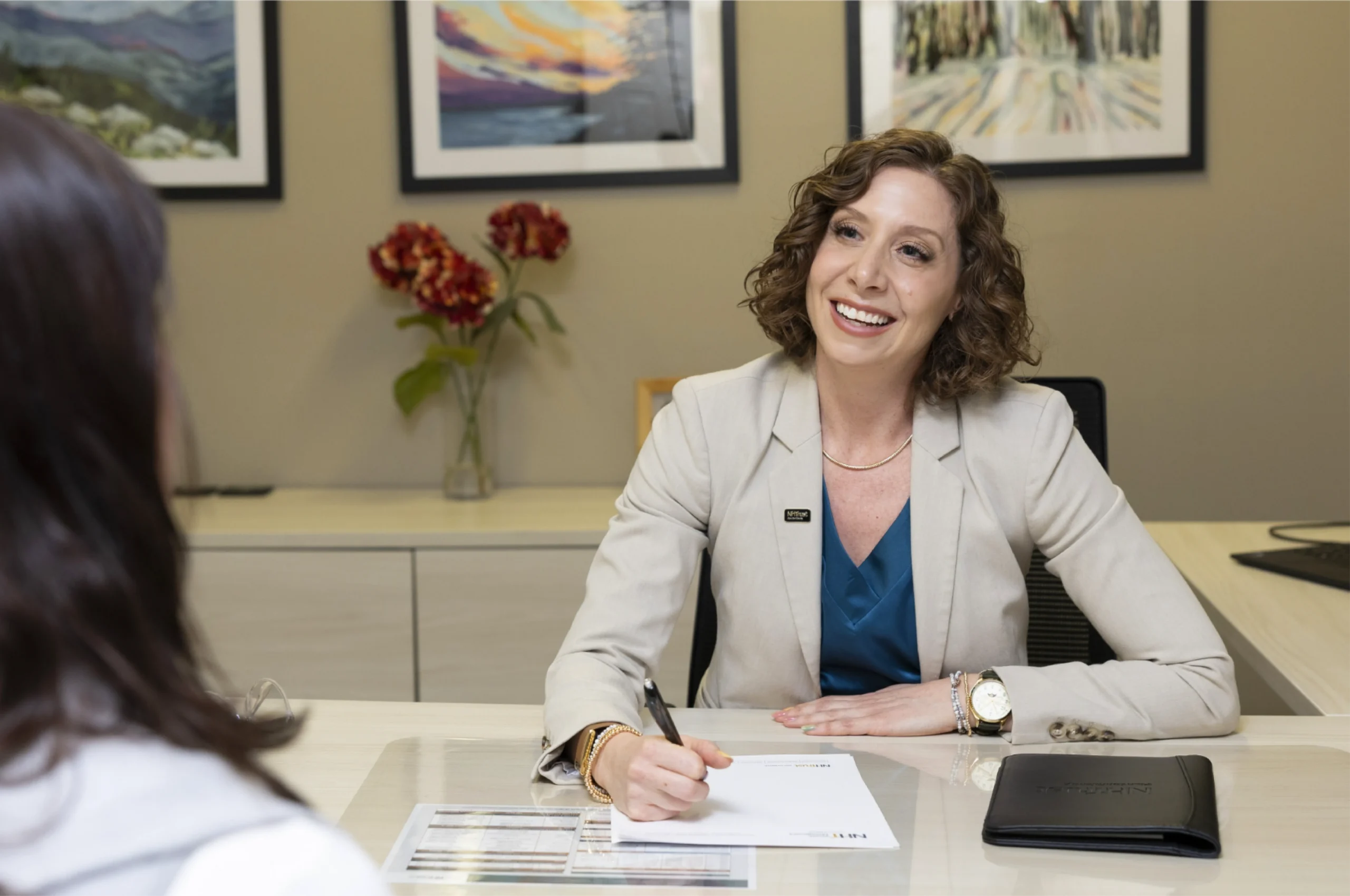 Smiling woman in a blazer talks to another person at a desk with papers and a tablet.