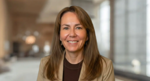 Woman with long brown hair in a tan blazer smiles in a softly lit, blurred indoor setting.