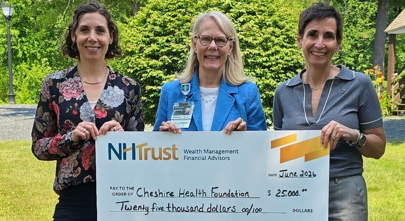 Three women hold a $25,000 NHTrust check for Cheshire Health Foundation outdoors, smiling at the camera.