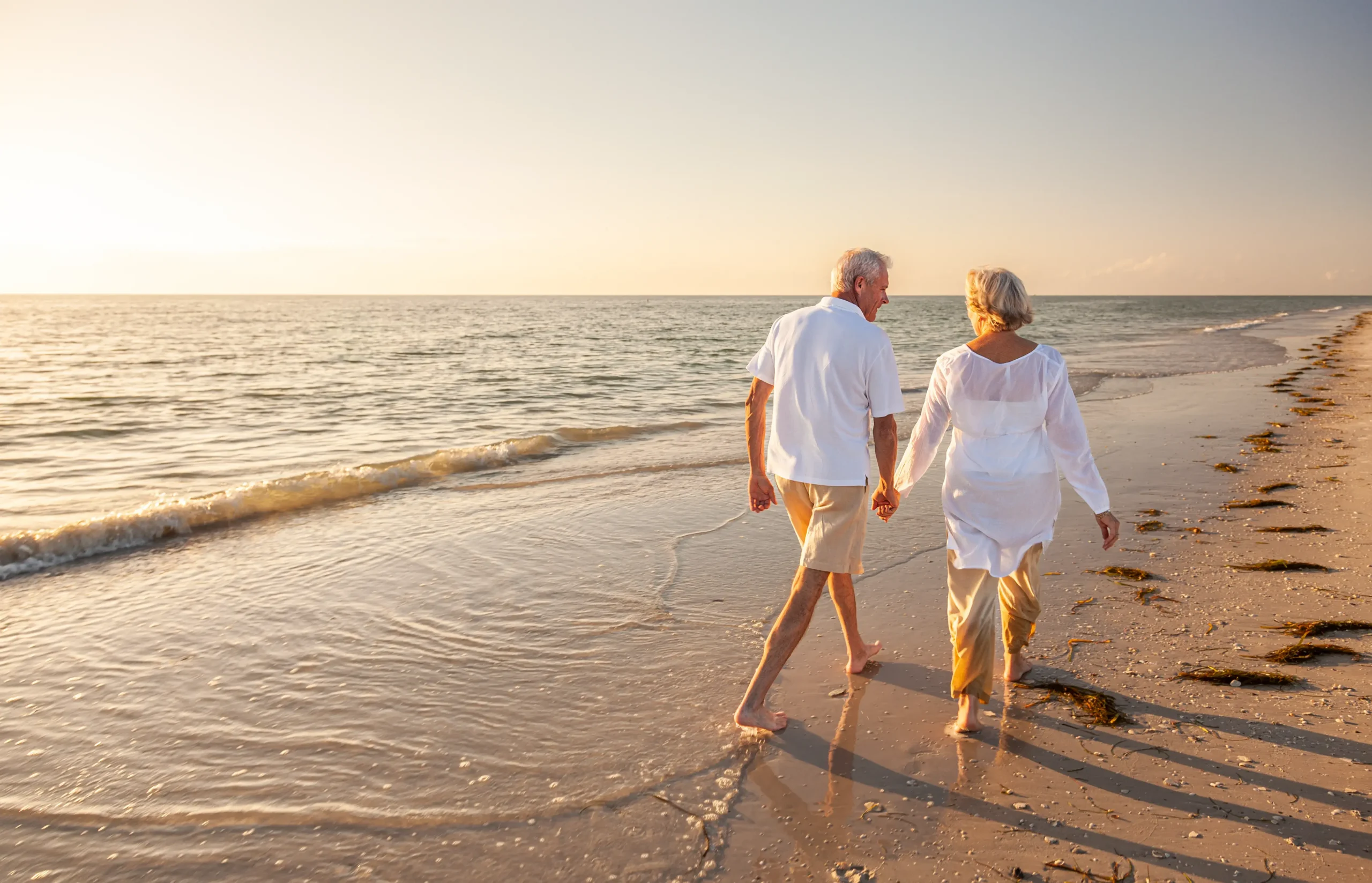 Retired older couple holding hands, walking barefoot along the beach at sunset, dressed in light clothing.