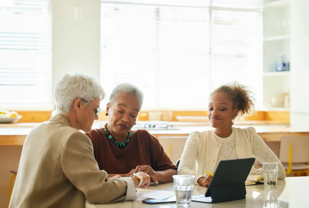 Three women sit at a table, discussing documents and using a laptop in a bright room.