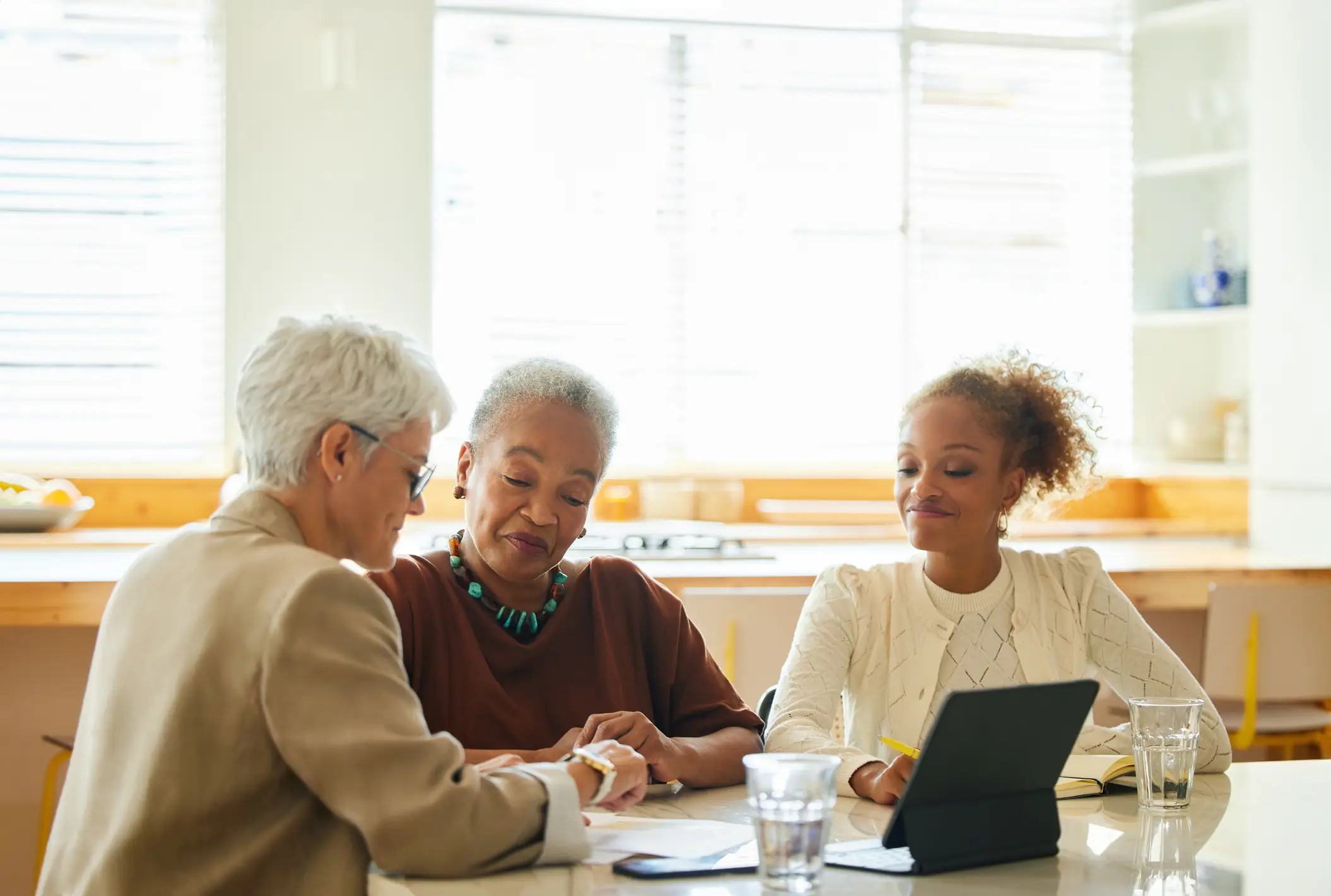 Three women sit at a table, discussing documents and using a laptop in a bright room.