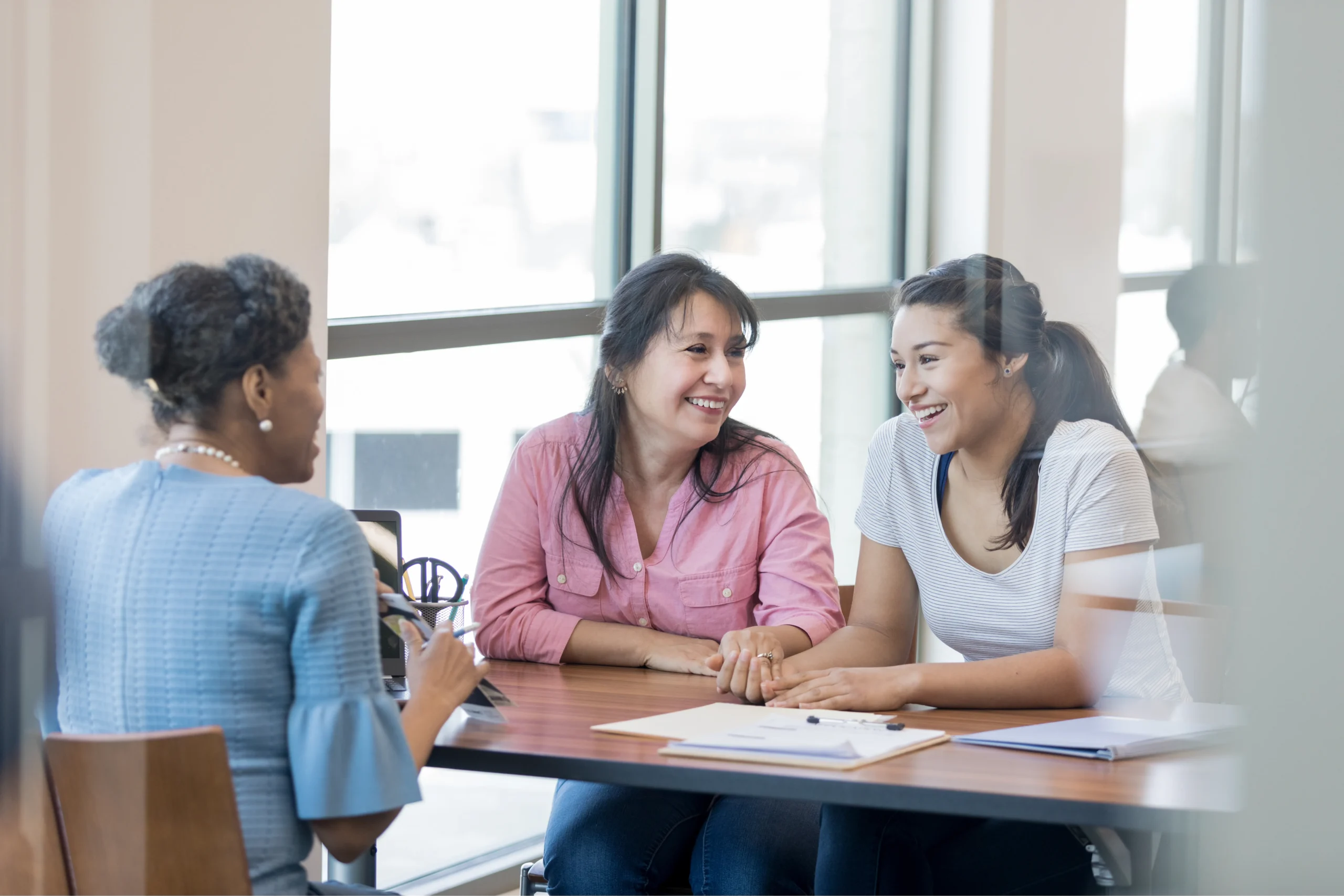Three women sit at a table by a window, talking and smiling with notebooks and papers in front of them.