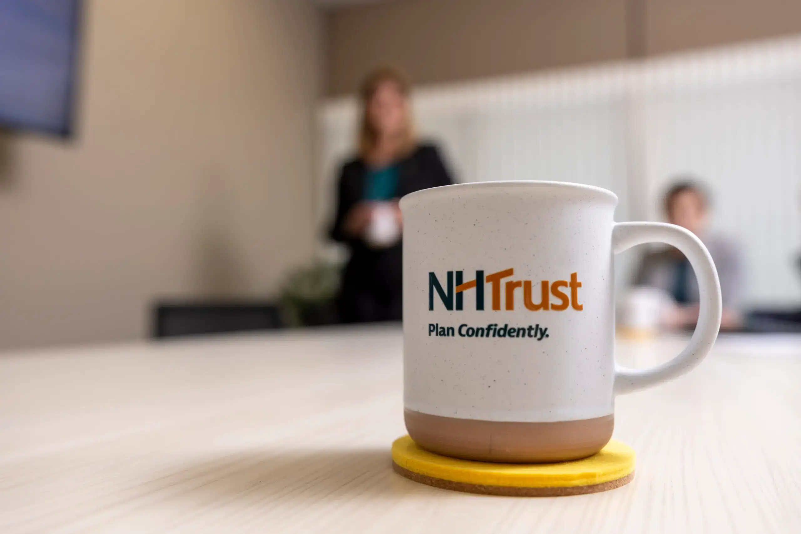 A ceramic mug featuring the NHTrust logo and the text Plan Confidently sits on a yellow coaster.