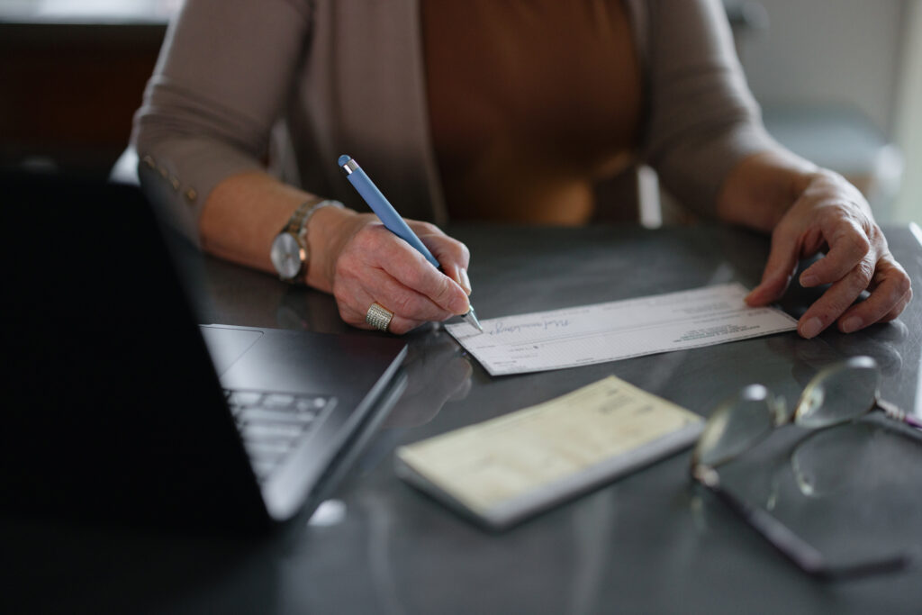 A person sitting at a desk signs a personal check with a blue pen next to a laptop and a checkbook.
